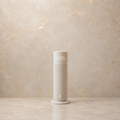 A minimalist cylindrical electric diffuser in a light beige textured finish, standing on a matching circular base against a textured beige marble wall. The top of the diffuser features the monogram 'AMS'.