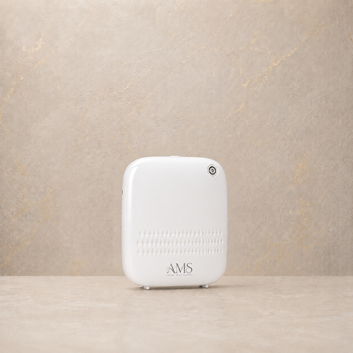 A white diffuser with the brand name 'AMS' printed on the front, standing on a light beige surface against a textured beige wall.