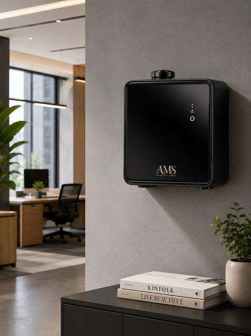 A black wall-mounted AMS diffuser with gold lettering, shown mounted on a gray wall in a modern office setting with a desk, chair, and books below.