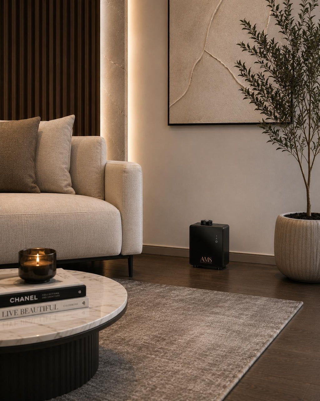 A modern living room scene featuring a AMS diffuser, light beige textured sofa, a round marble-top coffee table with a candle and a book, a gray area rug, a black speaker, a potted plant, and a dark wood slatted accent wall.