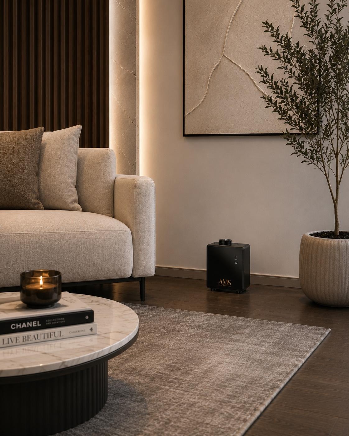 A modern living room scene featuring a AMS diffuser, light beige textured sofa, a round marble-top coffee table with a candle and a book, a gray area rug, a black speaker, a potted plant, and a dark wood slatted accent wall.