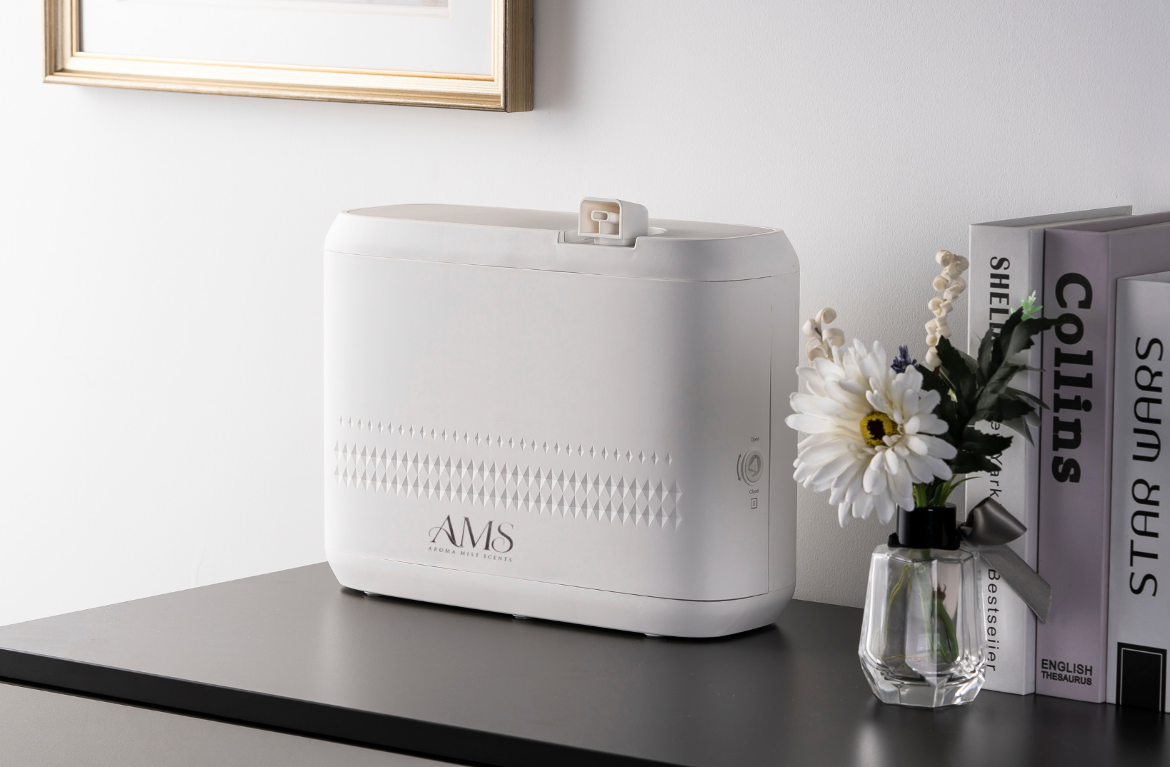A white diffuser with the brand name 'AMS' printed on the front, styled on a dark tabletop next to a small glass vase with white flowers and books against a light gray wall.