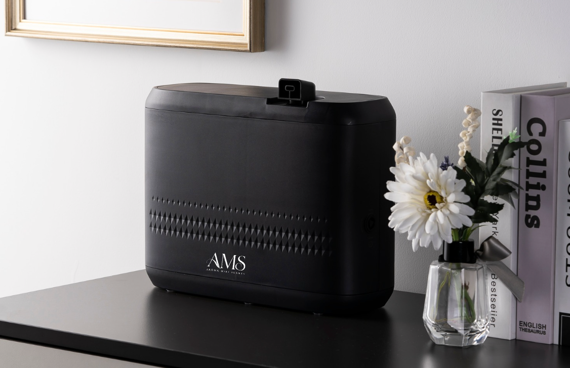 A black diffuser with the brand name 'AMS' printed on the front, sitting on a dark tabletop next to a small glass vase with white flowers and books against a light gray wall.