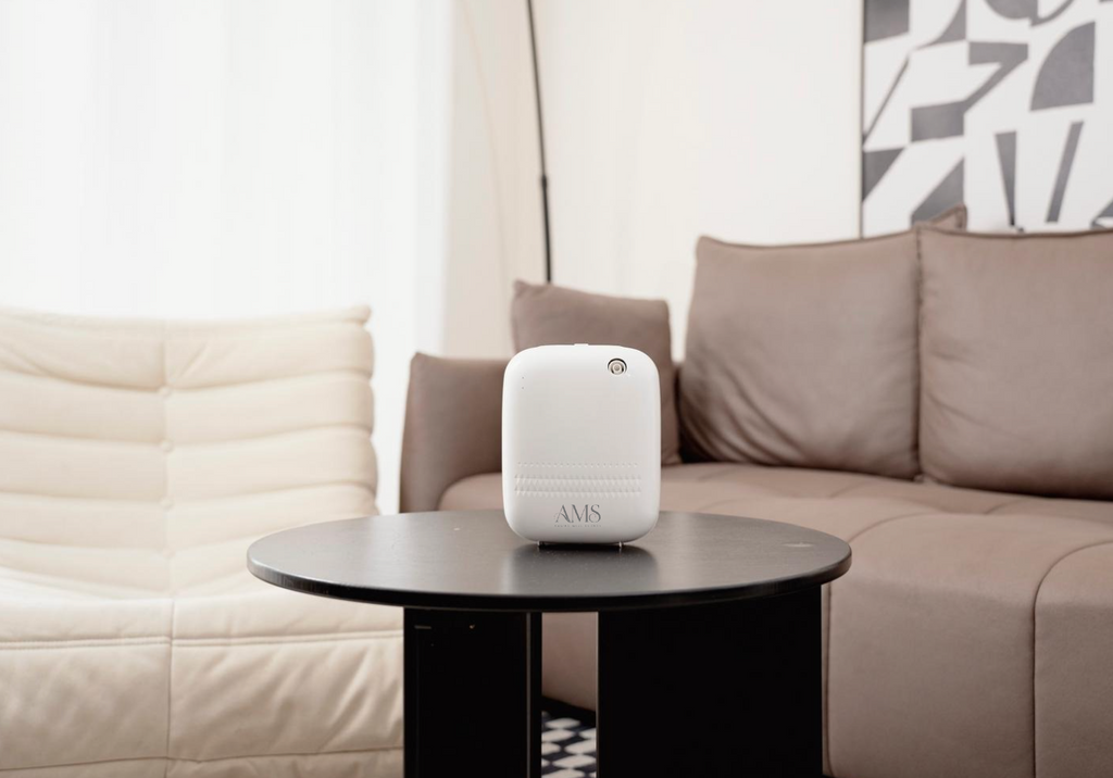 A white cylindrical smart speaker or humidifier with a small circular logo on top and a monogram 'MVS' at the bottom, displayed on a black round coffee table in a modern living room setting.
