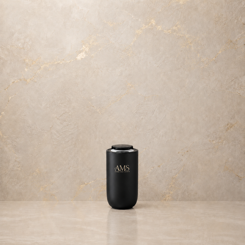 A matte black cylindrical diffuser with a gold 'AMS' logo, standing on a polished stone surface against a textured beige marble wall.