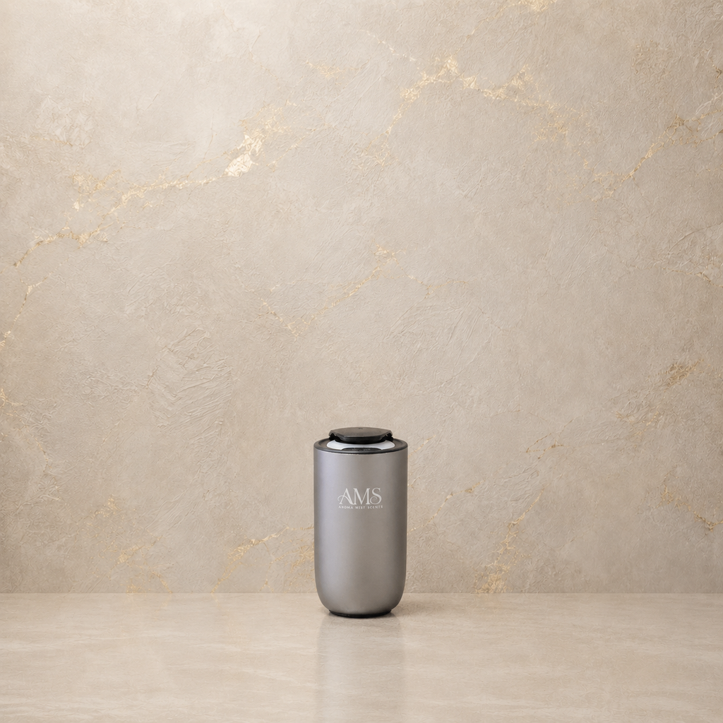 A sleek, matte gray cylindrical diffuser with a black lid and the monogram 'AMS' printed on the front, set on a light beige stone surface against a textured beige marble wall.