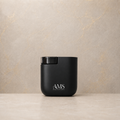 A matte black cylindrical diffuser with a clear top window and white 'AMS AESTHETICS MART' branding, set on a light beige stone surface against a textured beige wall.