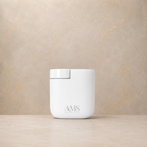 A white cylindrical diffuser with a matching white lid sits on a light beige surface against a textured beige wall. The front of the container is printed with the brand name 'AMS'