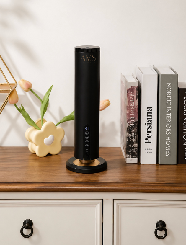 A sleek black cylindrical AMS diffuser with a gold accent base, displayed on a wooden cabinet next to decorative books and a flower vase.