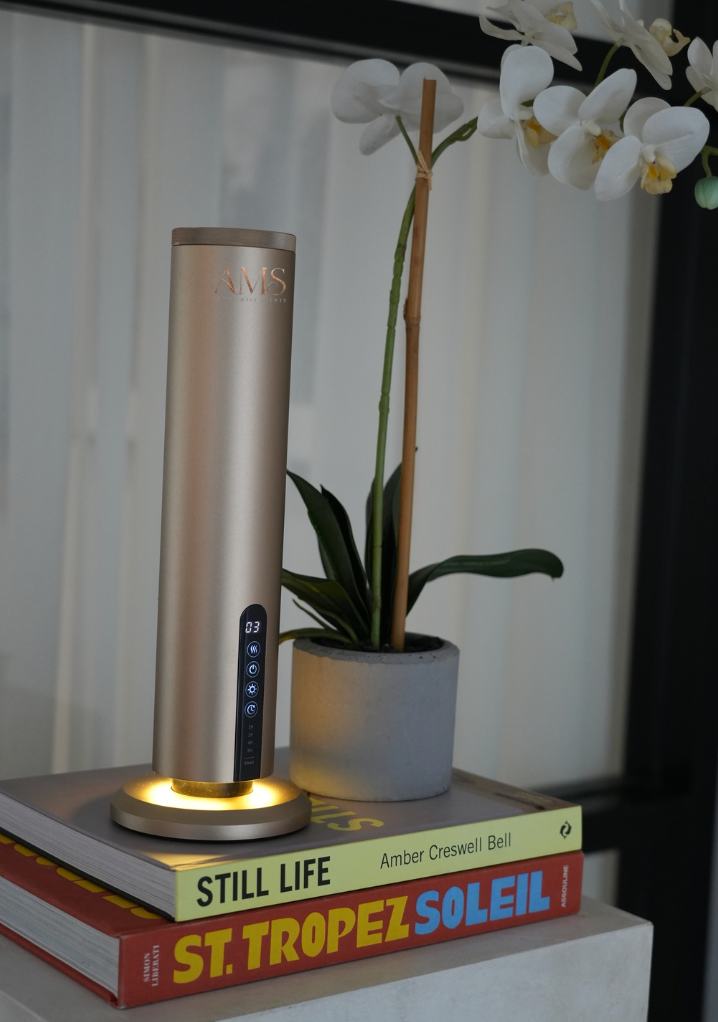 A sleek, metallic gold cylindrical diffuser with a glowing warm light base stands on top of stacked books next to a potted white orchid.