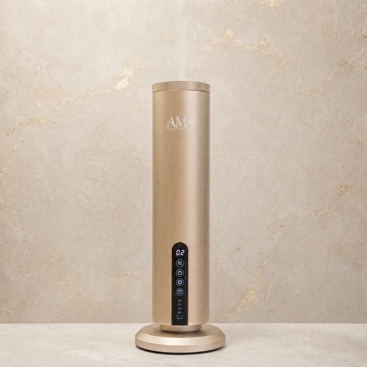 A tall cylindrical AMS diffuser in a brushed champagne gold finish, standing on a matching round base against a textured beige stone wall. A digital display on the front shows the temperature and humidity levels.