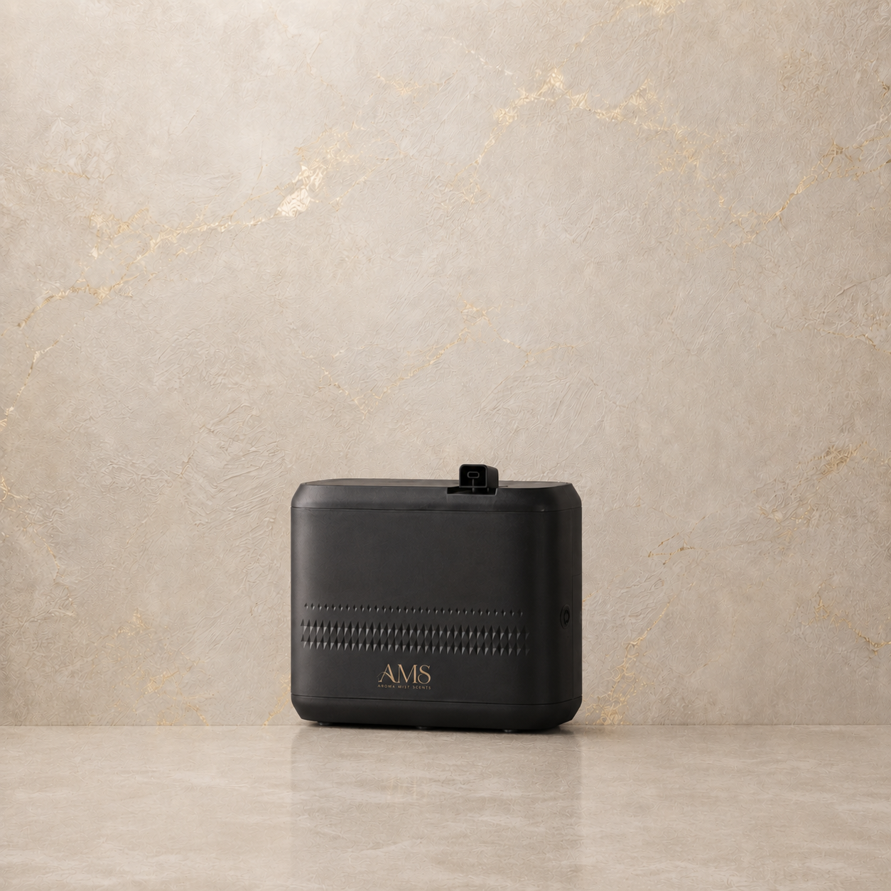 A black rectangular electronic diffuser with the brand name 'AMS' in gold lettering, resting on a light beige stone surface against a matching stone wall.