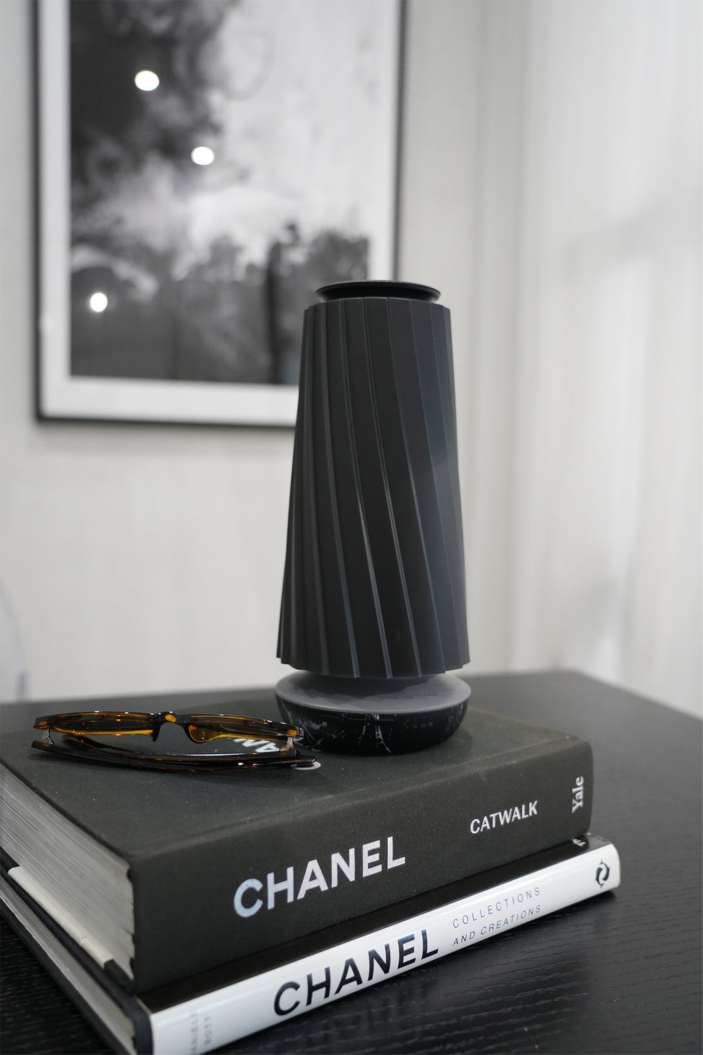 A dark gray pleated cylindrical diffuser sits on top of two stacked Chanel books on a dark table, with a pair of sunglasses resting on the top book. A blurred black and white framed print hangs on the wall in the background.
