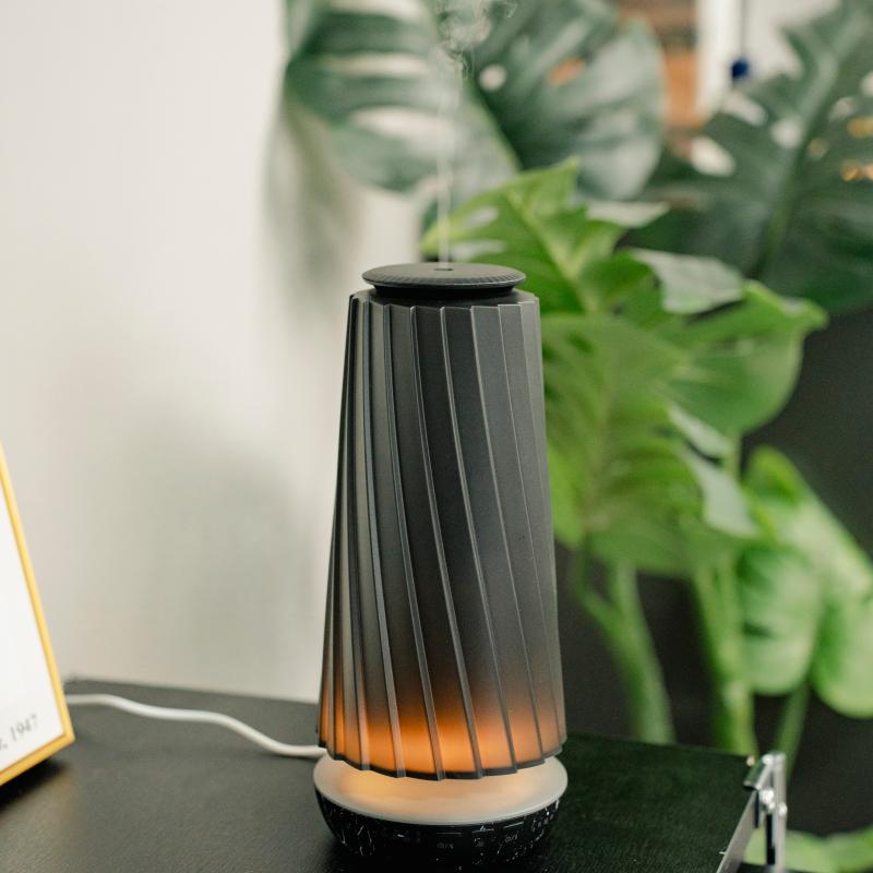 A modern dark gray pleated diffuser with a warm orange glow at the base, emitting a fine mist from the top, set on a dark tabletop with green plants in the background.