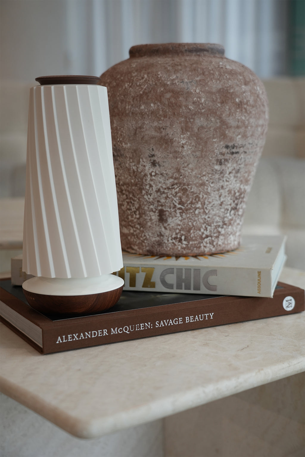 A modern white ribbed diffuser with dark brown wooden accents sits on a stack of books next to a large textured brown ceramic vase on a marble shelf.