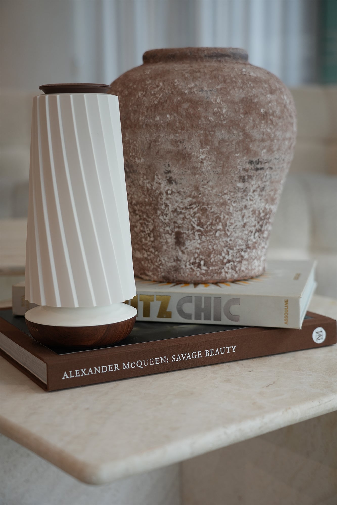 A modern white ribbed diffuser with dark brown wooden accents sits on a stack of books next to a large textured brown ceramic vase on a marble shelf.