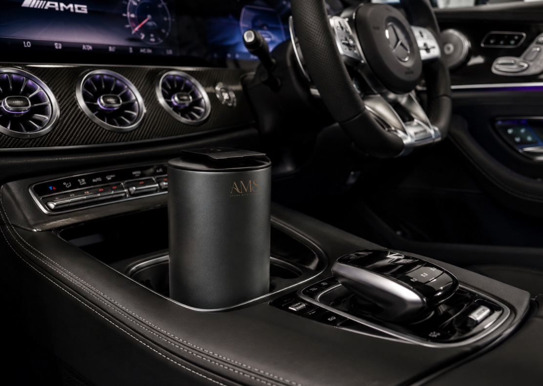 A black cylindrical AMS portable diffuser with a brushed metal top sits in a luxury car center console. The background shows a black leather interior with carbon fiber trim, a Mercedes-Benz steering wheel, and an AMG-branded dashboard.