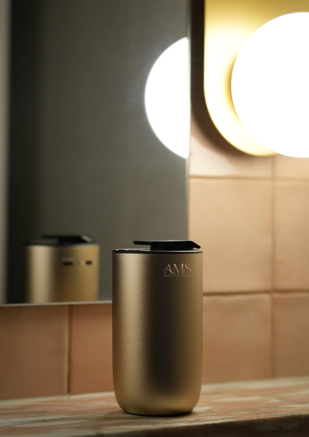 A matte metallic gold cylindrical diffuser with a black lid sits on a wooden counter in a warm, modern bathroom setting with a mirror reflection.