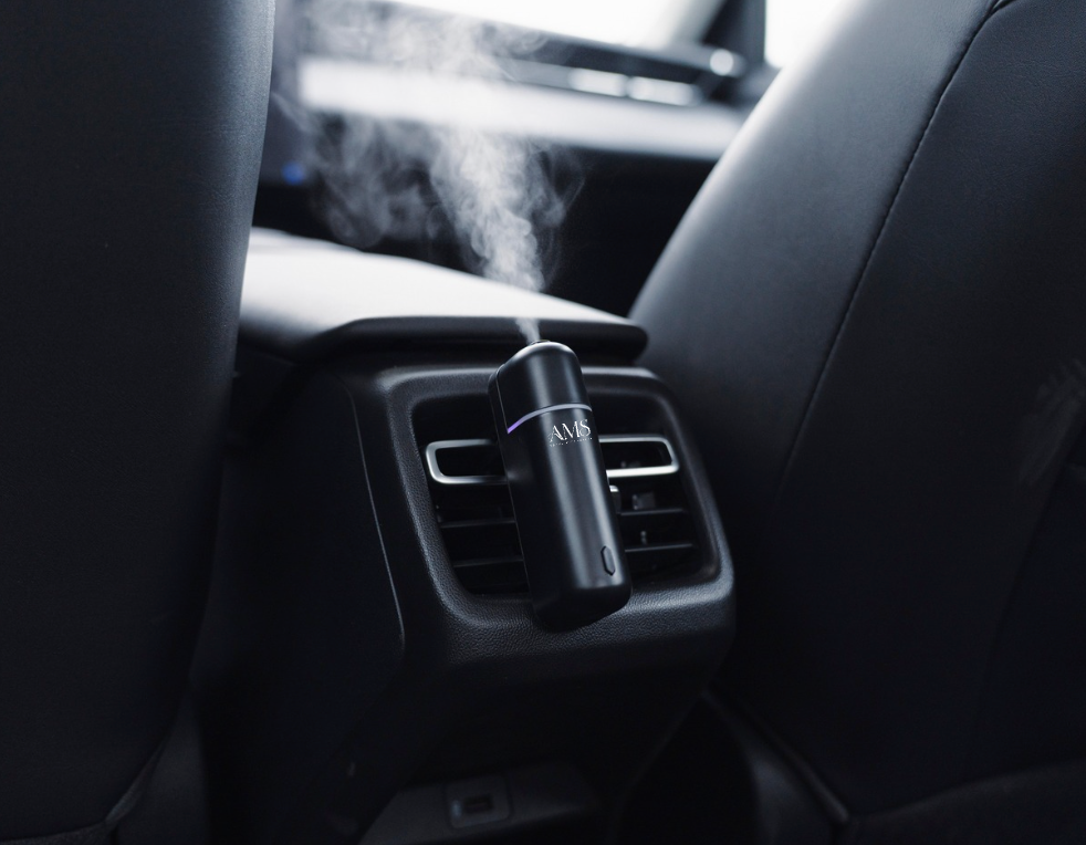 A black car diffuser with visible mist emitting from the top, clipped onto a car air vent between the rear seats.