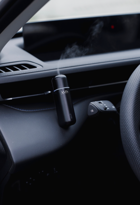 A black cylindrical car diffuser with the brand name 'AMS' printed in white, shown plugged into a car's dashboard air vent with a plume of mist rising.