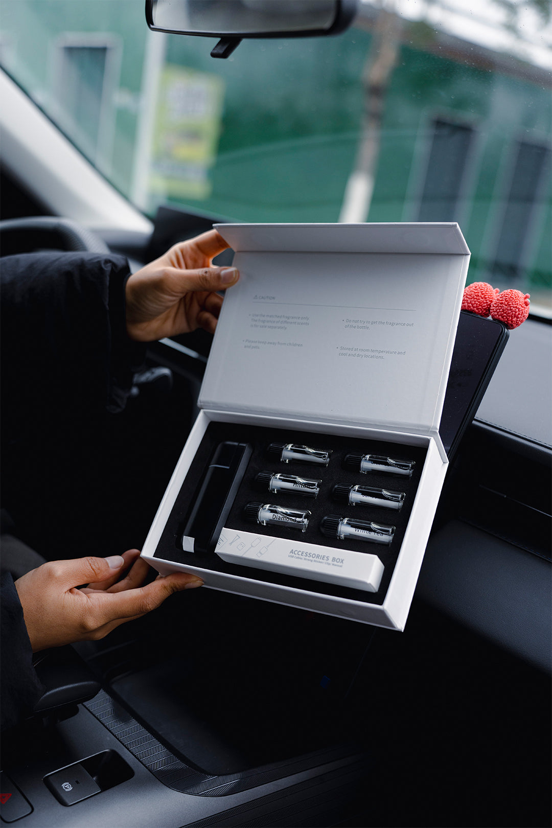 A person holding an open white gift box containing several small fragrance vials inside a car interior.
